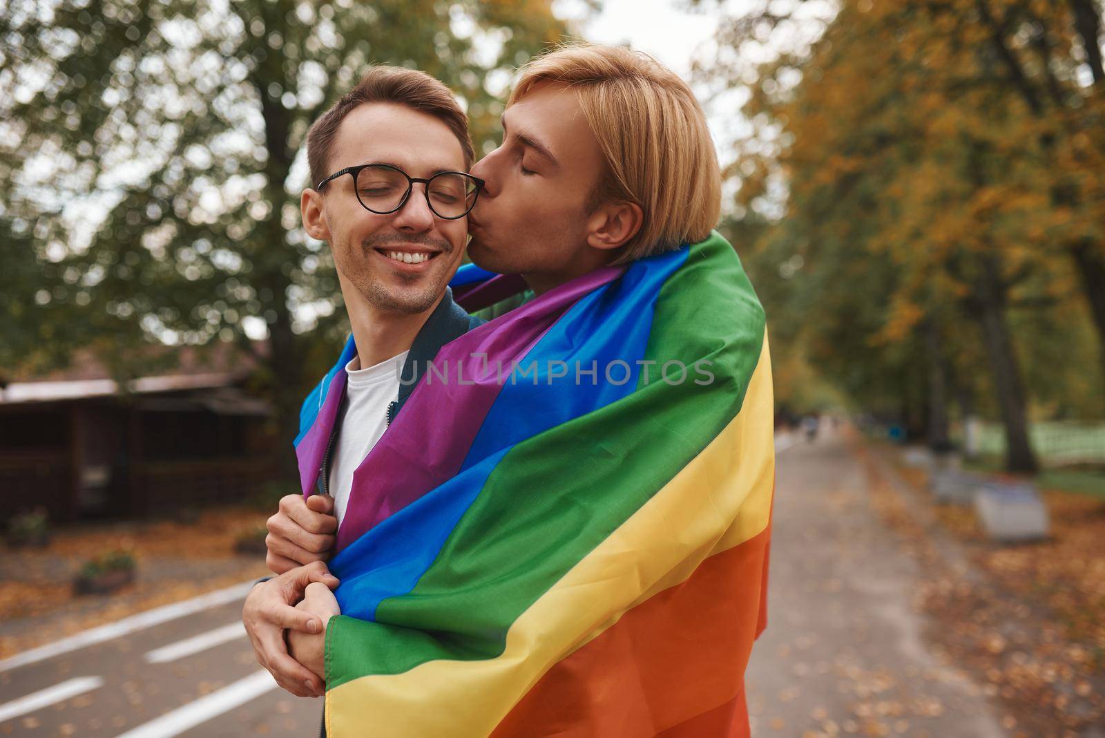 Romantic gay couple hugging, kissing and holding hands outdoors. Two handsome men holding LGBT pride flag in park.