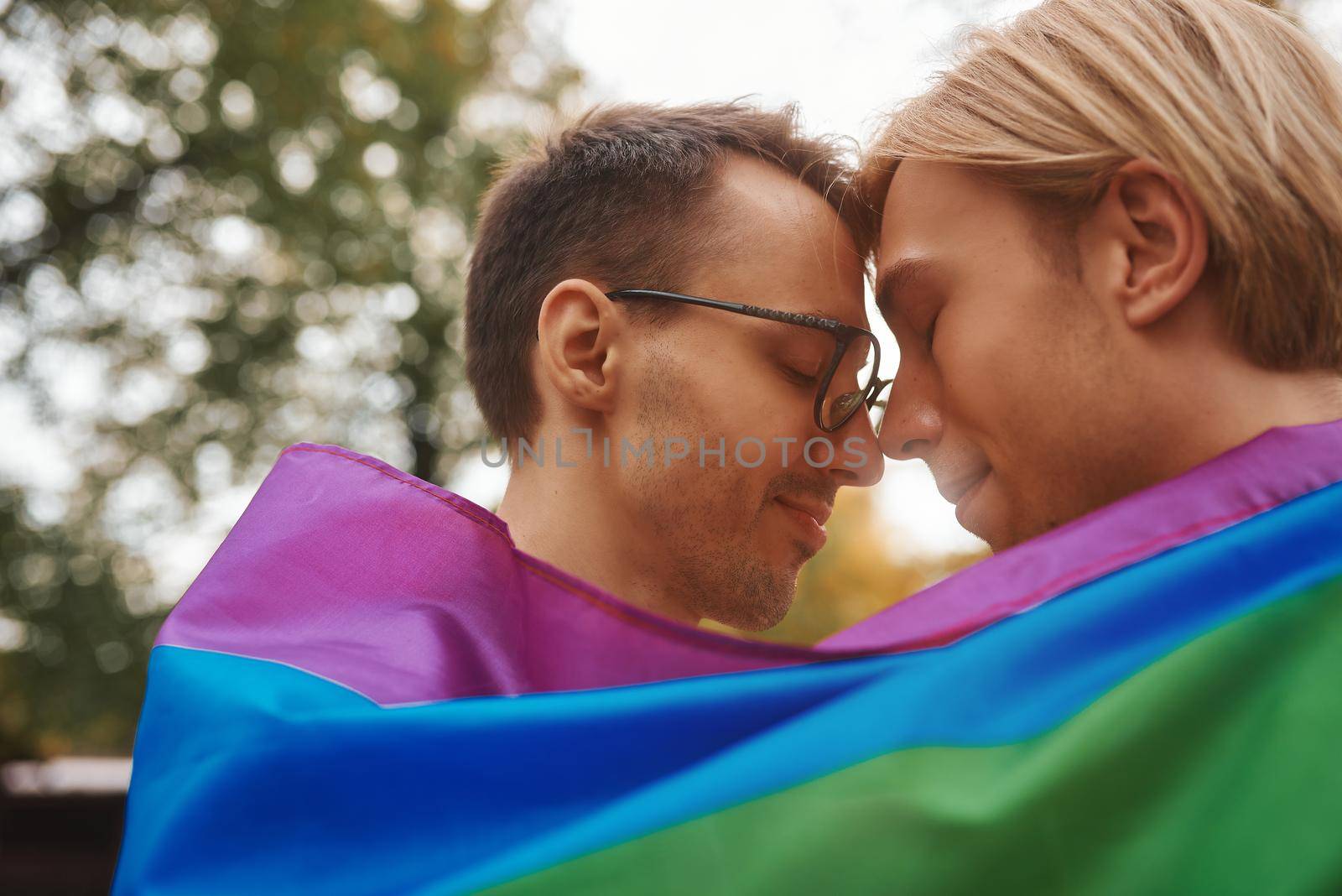 Romantic gay couple hugging, kissing and holding hands outdoors. Two handsome men holding LGBT pride flag in park.