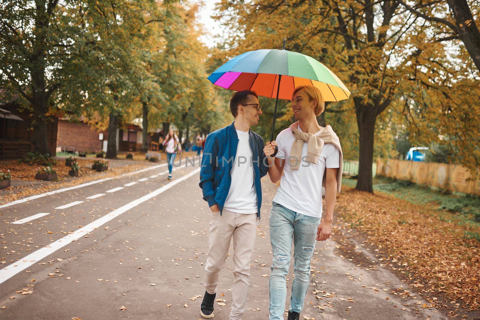Loving gay couple walking outdoors with rainbow umbrella. Two handsome men having romantic date in park. LGBT concept.