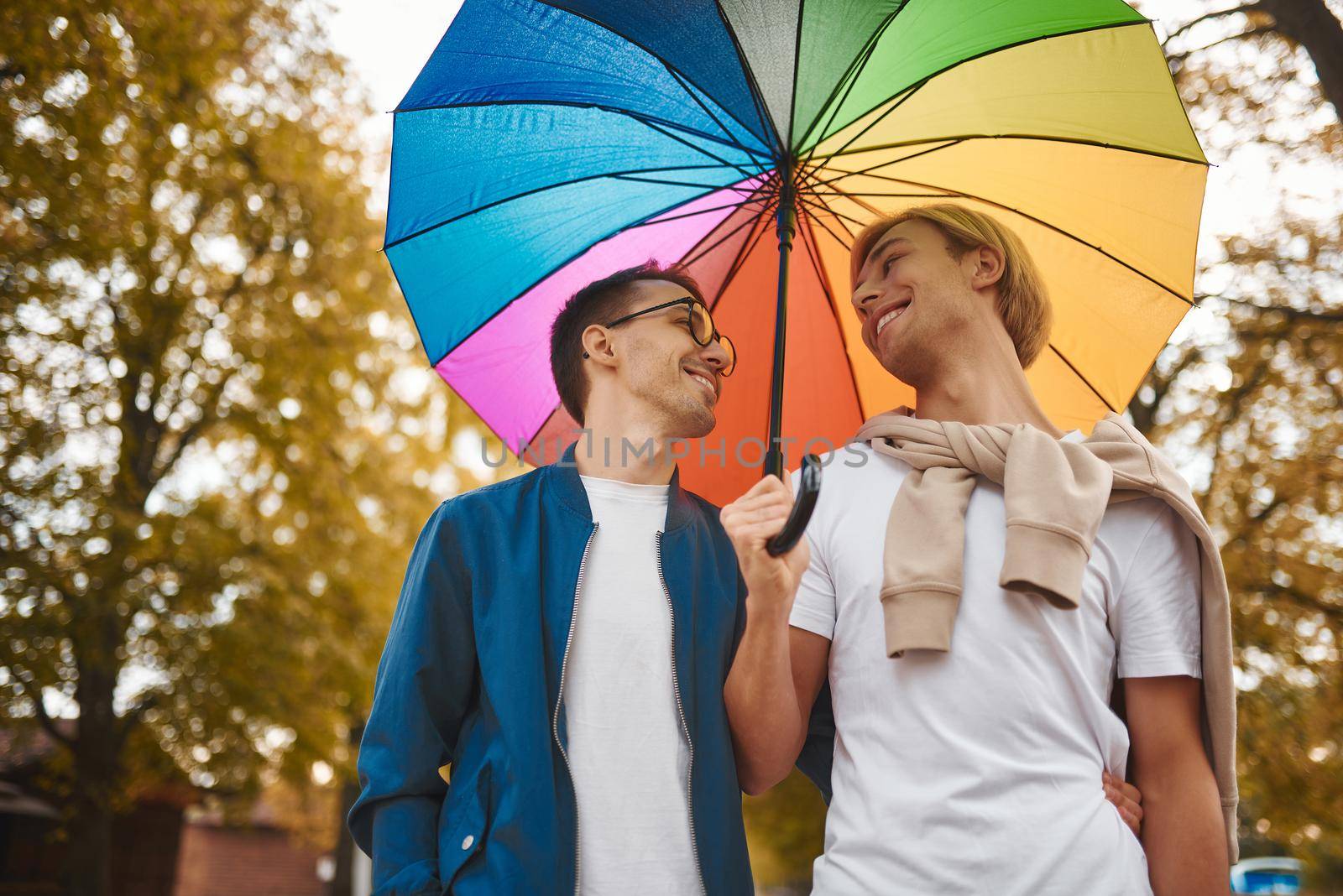 Loving gay couple walking outdoors with rainbow umbrella. Two handsome men having romantic date in park. LGBT concept.