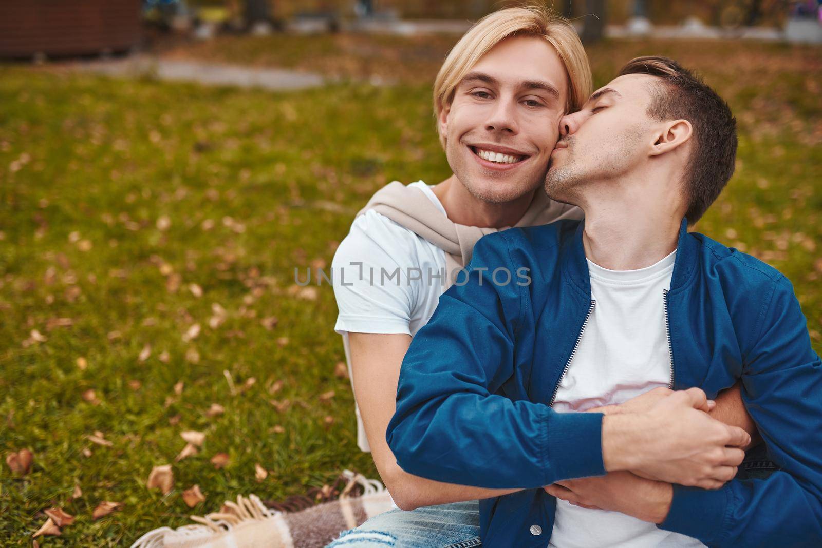 Loving gay couple having romantic date outdoors. Two handsome men sitting together on blanket in park and hugging. LGBT concept.