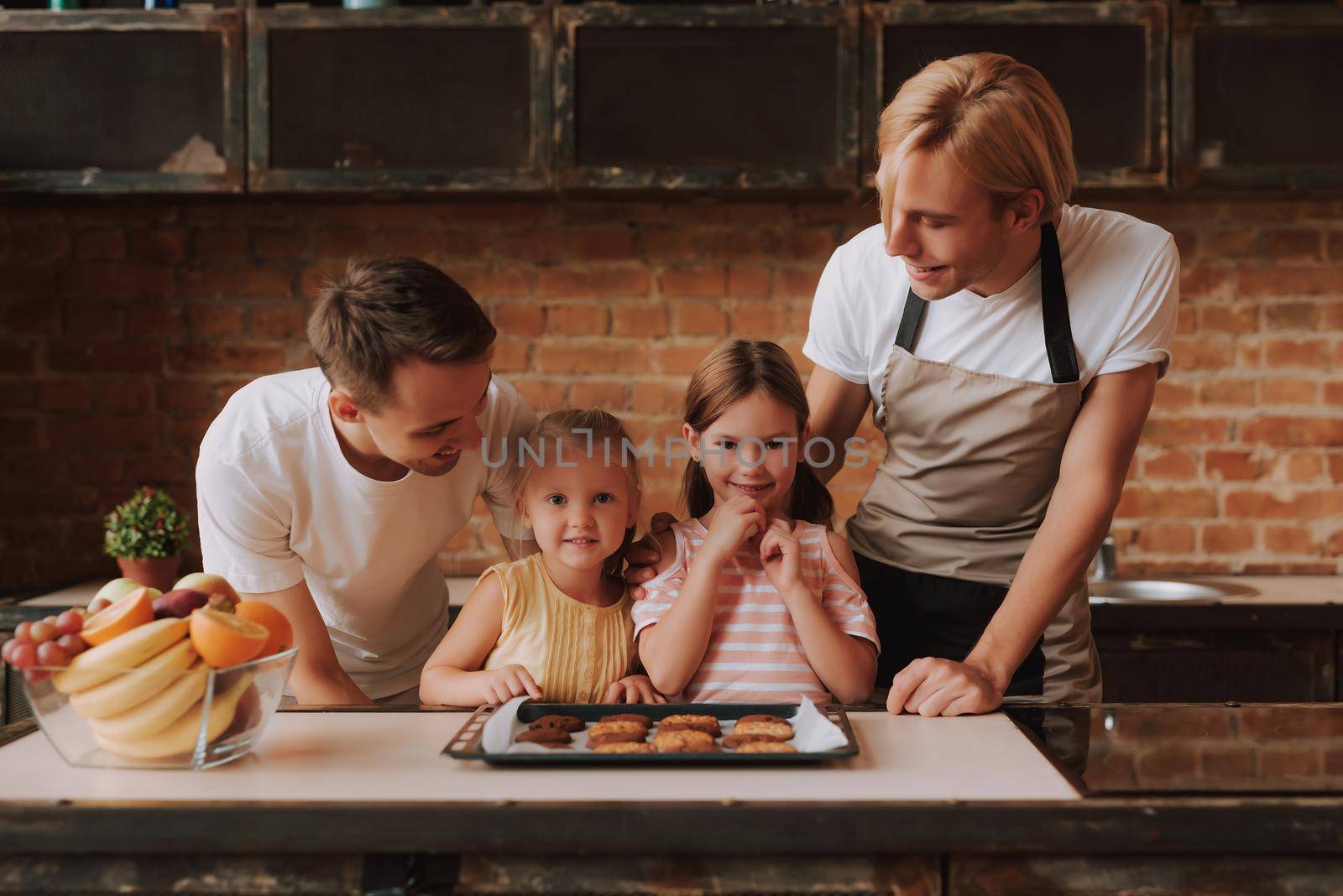 Gay couple with their adopted cute daughters cooking on kitchen. Lgbt family at home. Portrait of two handsome men and their two little daughters eating cookies.