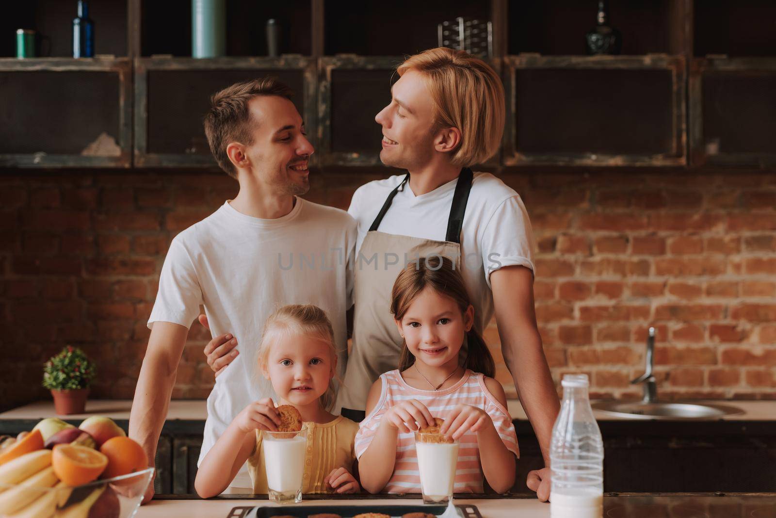 Gay couple with their adopted cute daughters cooking on kitchen. Lgbt family at home. Portrait of two handsome men and their two little daughters eating cookies and drinking milk.