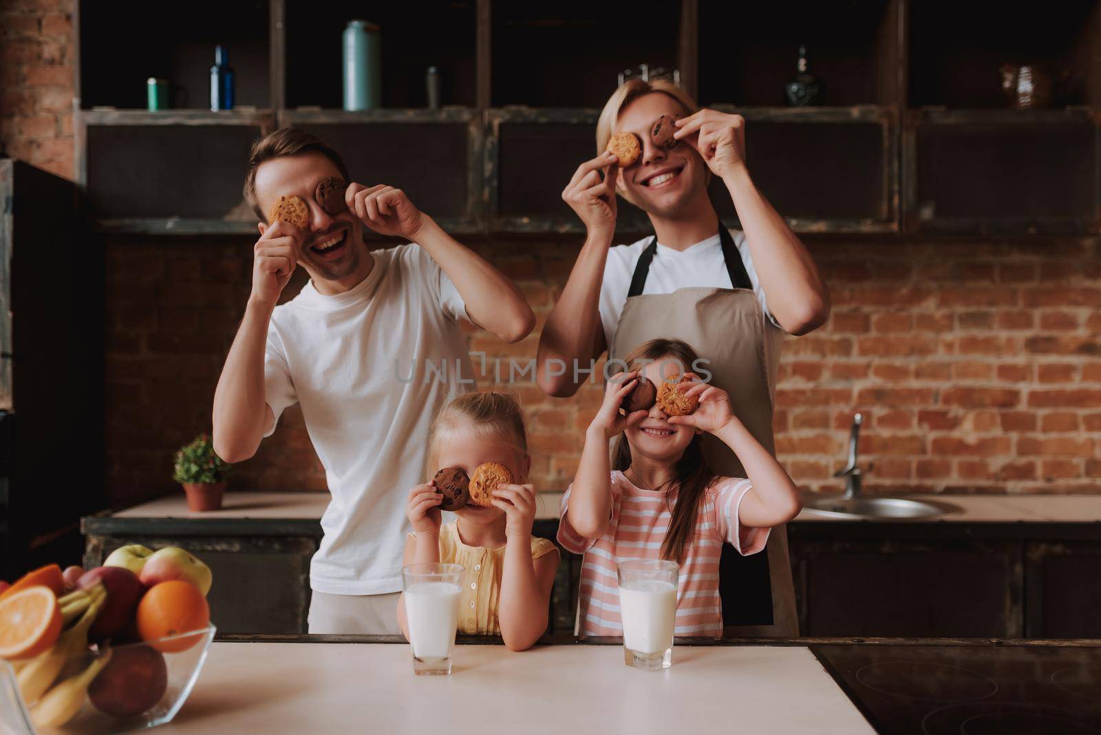 Gay couple with their adopted cute daughters cooking on kitchen. Lgbt family at home. Portrait of two handsome men and their two little daughters eating cookies and drinking milk.