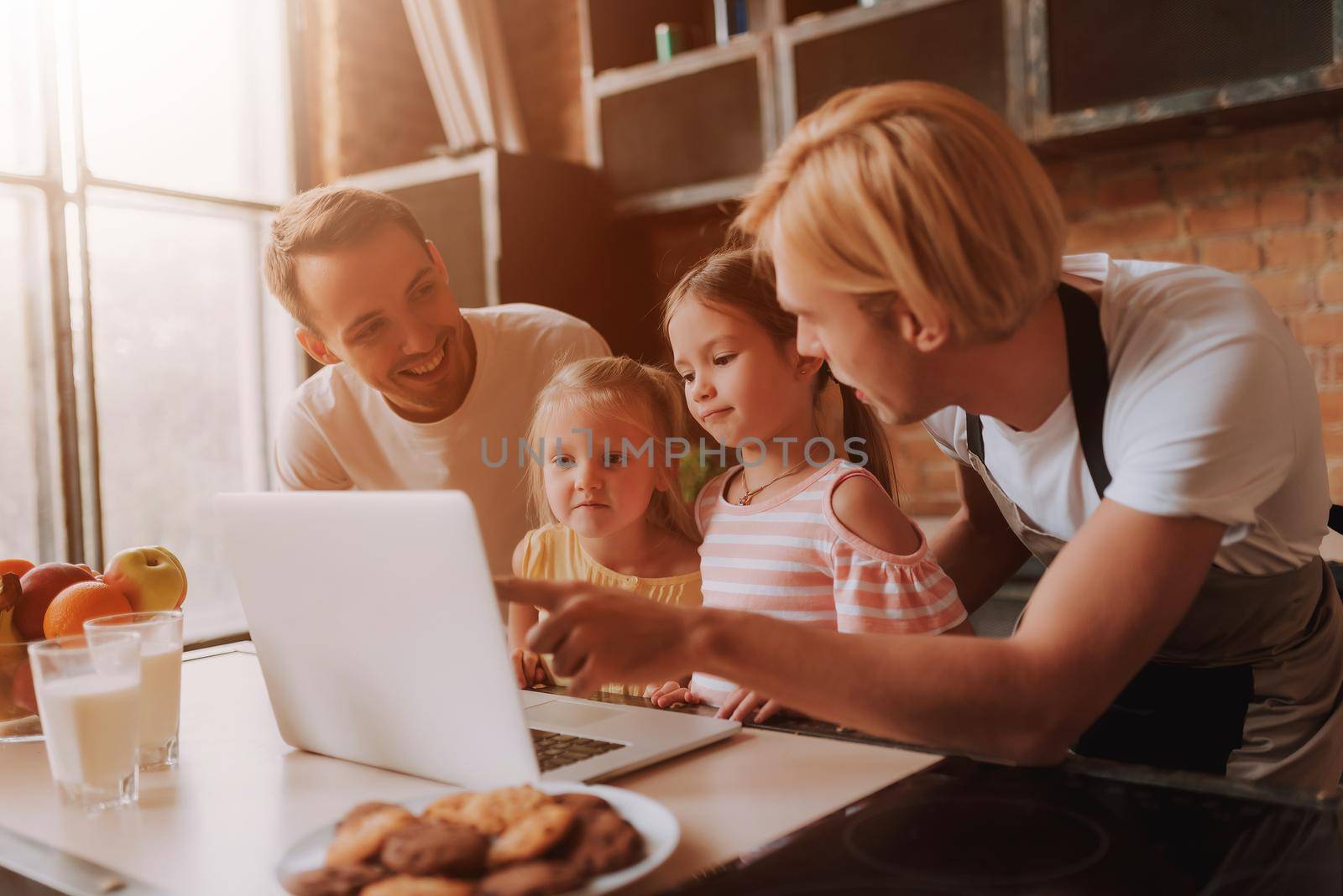 Gay couple with their adopted cute daughters reading recipe on laptop and cooking on kitchen. Lgbt family at home.