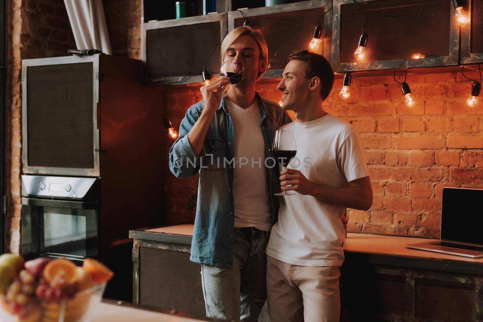 Loving gay couple at home. Two handsome men hugging and kissing on kitchen. Romantic atmosphere with bottle of red wine. LGBT concept.