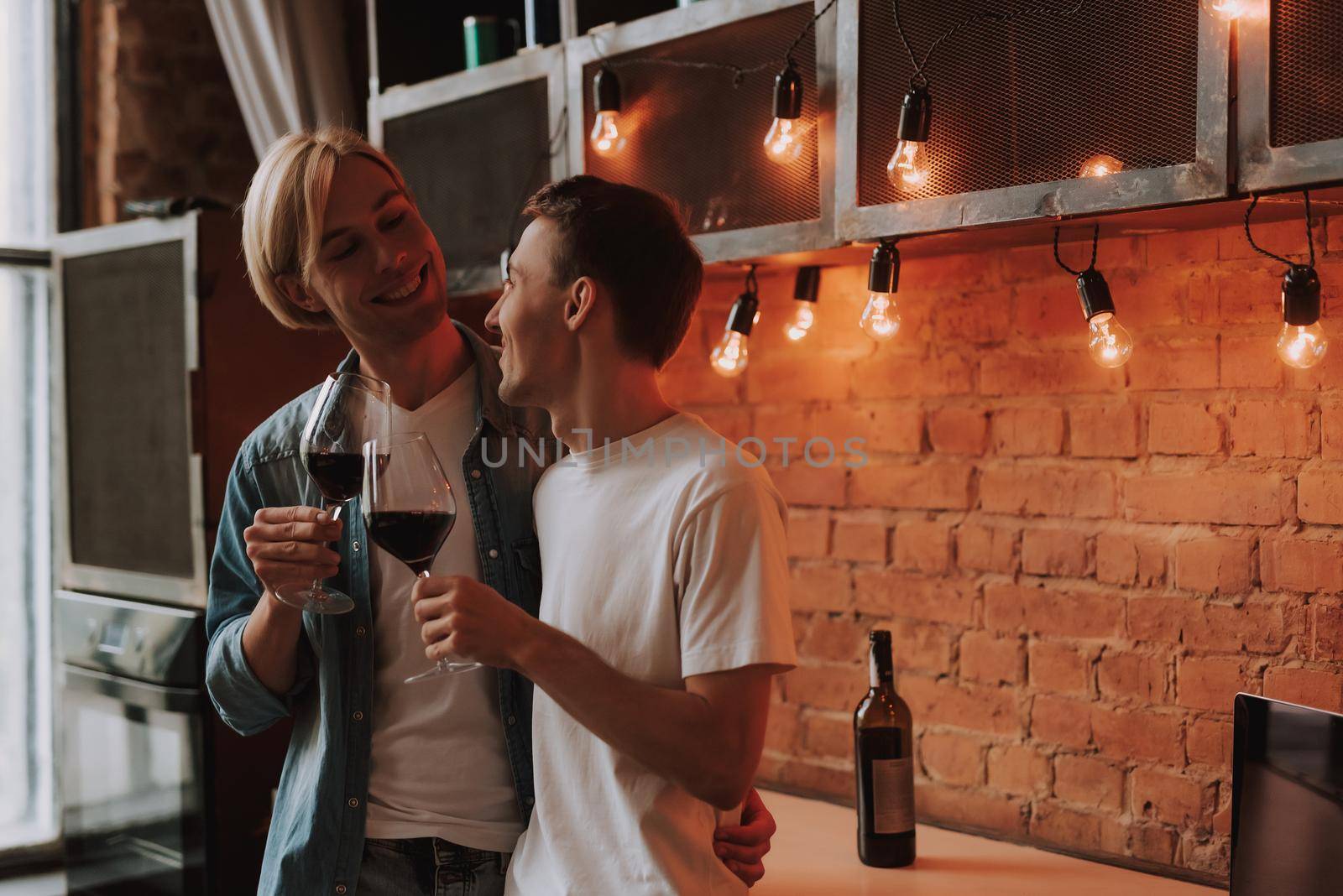 Loving gay couple at home. Two handsome men hugging and kissing on kitchen. Romantic atmosphere with bottle of red wine. LGBT concept.