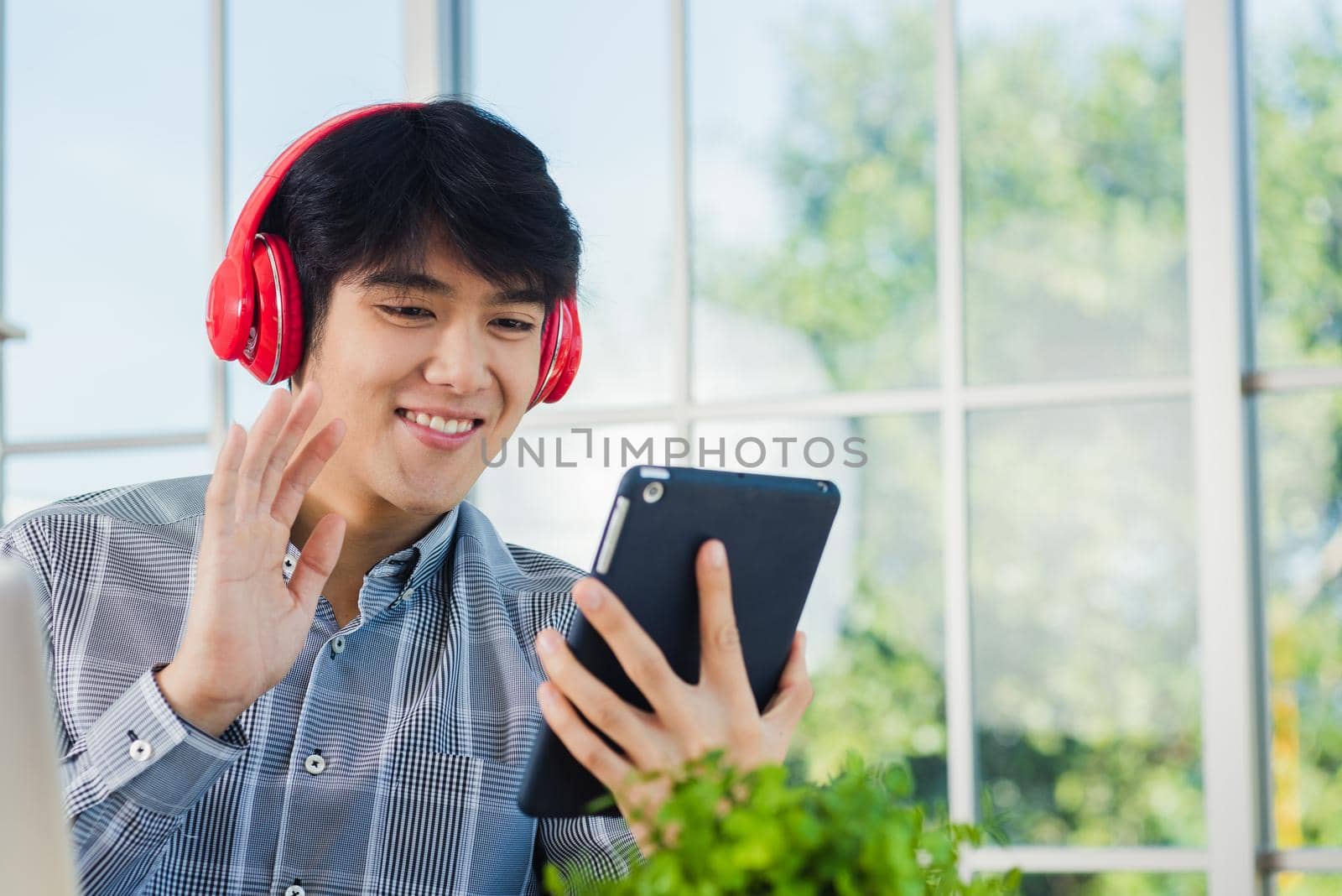 Asian young business man happiness with red headphones he sitting on desk workplace using digital tablet while greeting by video chat conferences talking and discussing