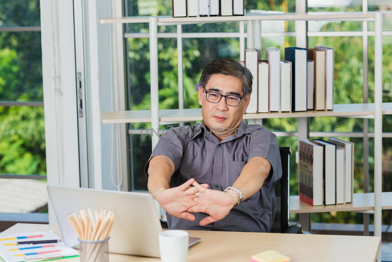 Asian businessman tired overworked he stretch oneself on the desk. senior man with eyeglasses break stretching his arms on table at his working place