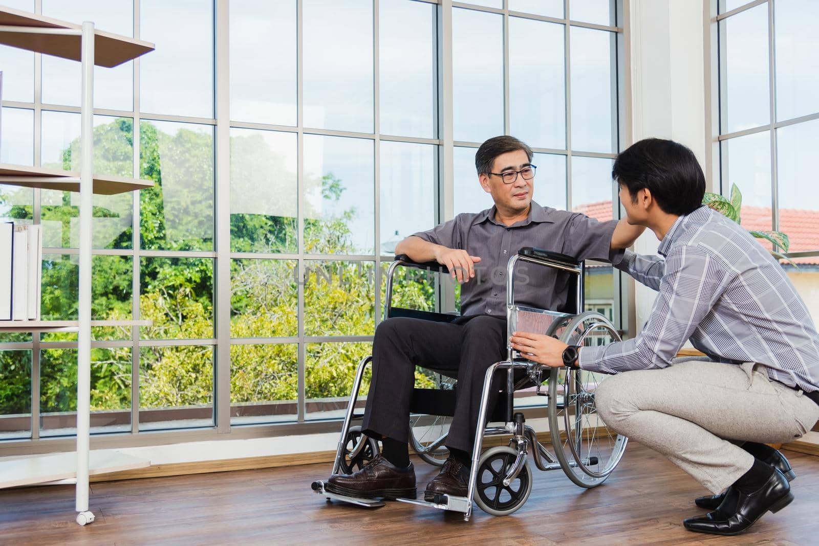 Asian senior disabled businessman in wheelchair discuss interacting together with the team in the office. The old man in a wheelchair and his young son talking to and comforting bound father