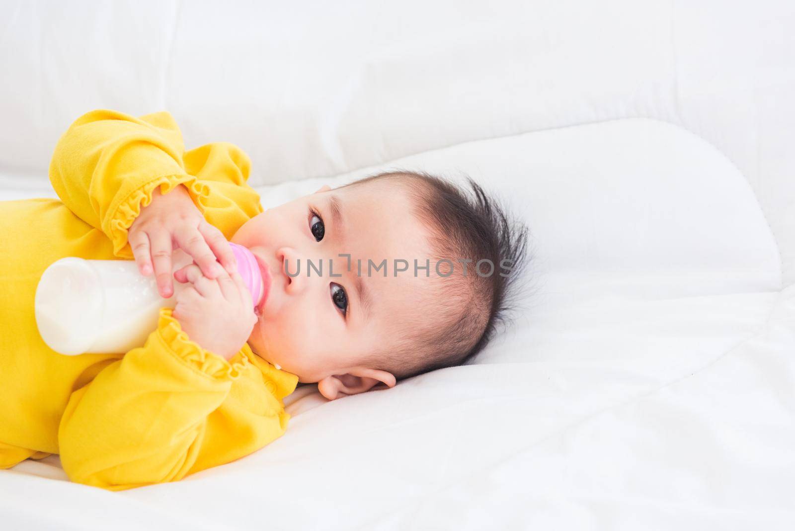 Asian beautiful little baby girl wearing a yellow dress eat milk sleeping feeding lying on the white bed, infant holding a bottle of milk, baby food concept