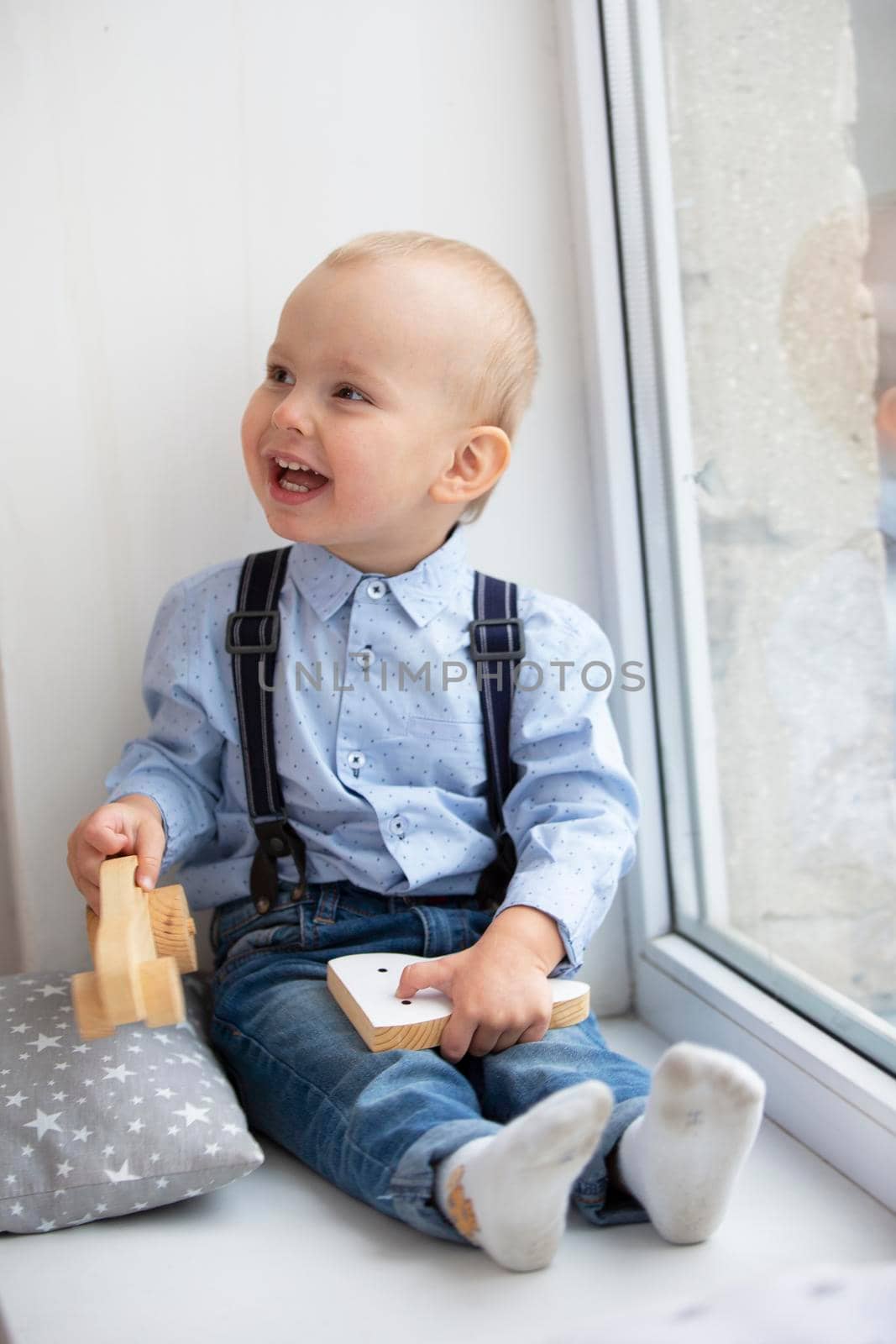 Little baby near the window. The cheerful kid with a toy sits on a window sill.
