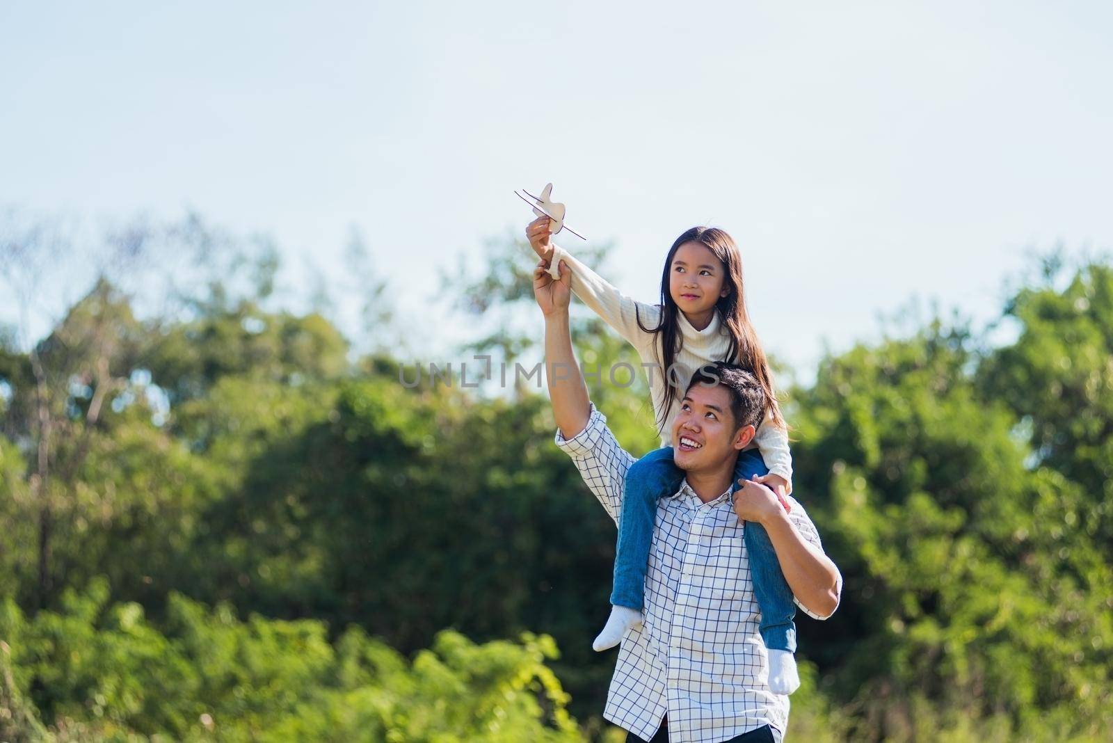 Happy Asian young family father and carrying an excited girl on shoulders having fun and enjoying outdoor lifestyle together playing aircraft toy on sunny summer day, Father's day concept