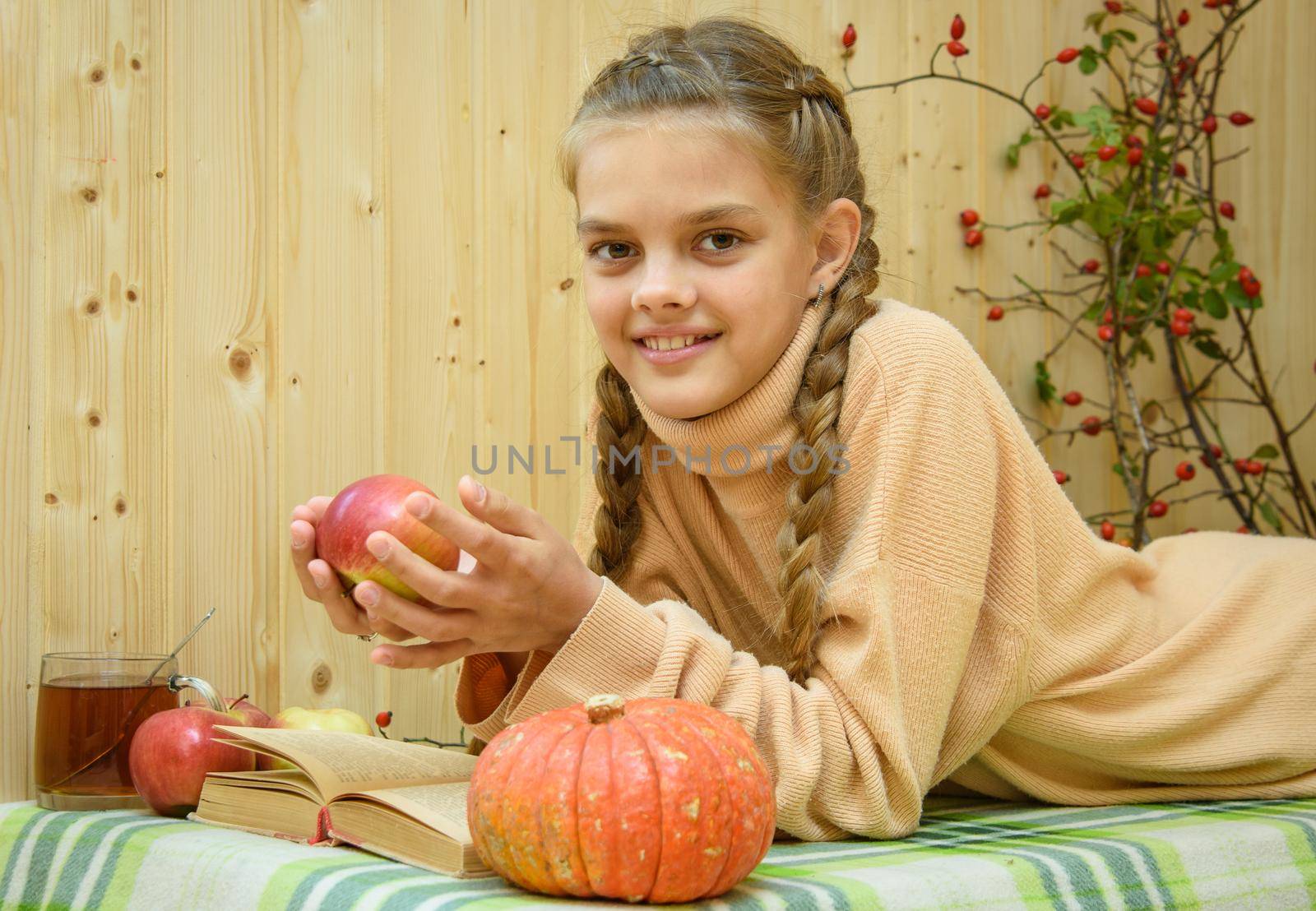A girl lying down reads a book, holds an apple in her hands and looks happily into the frame