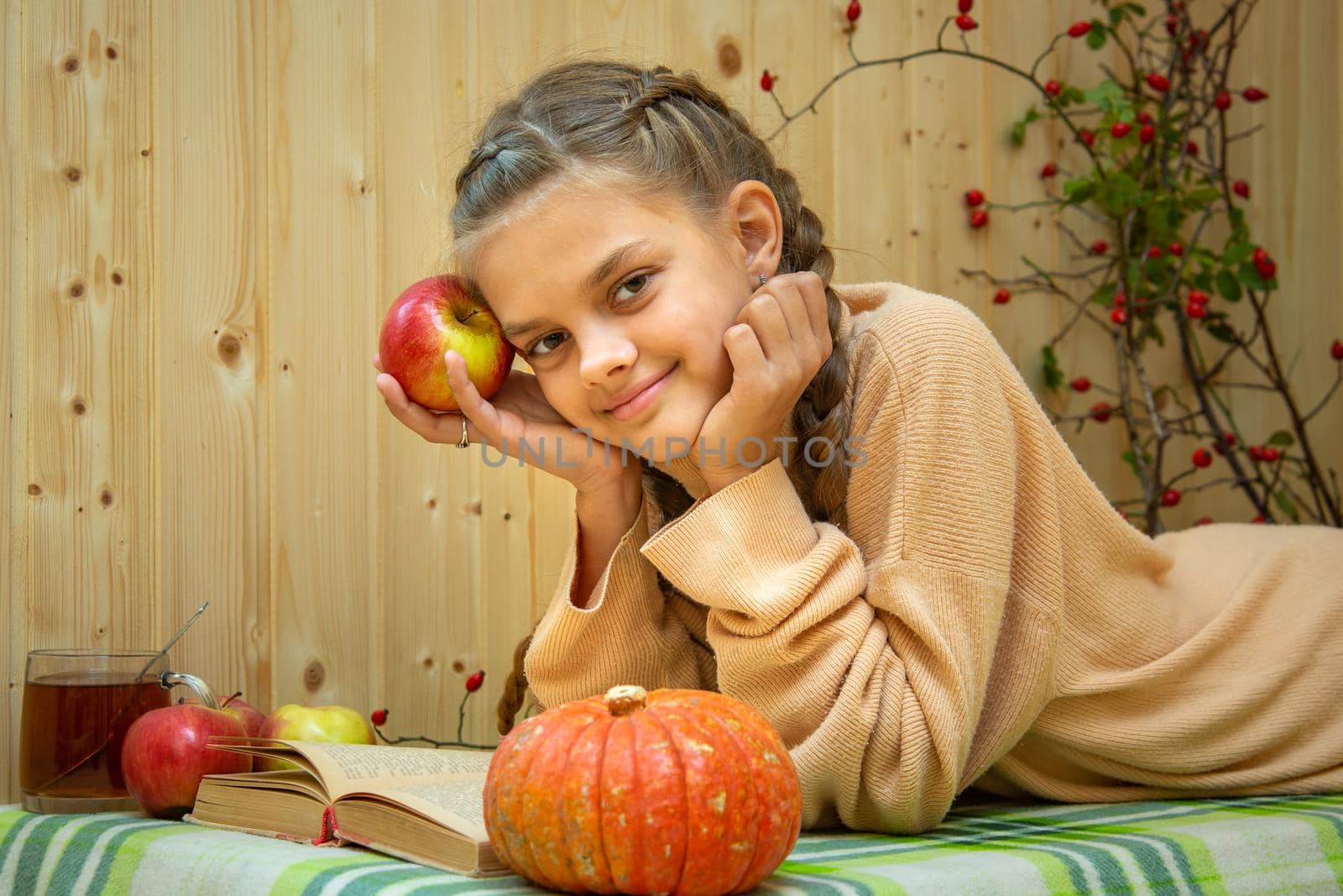 A girl lying down reads a book, a pumpkin and apples lie nearby, a girl joyfully looks into the frame by Madhourse