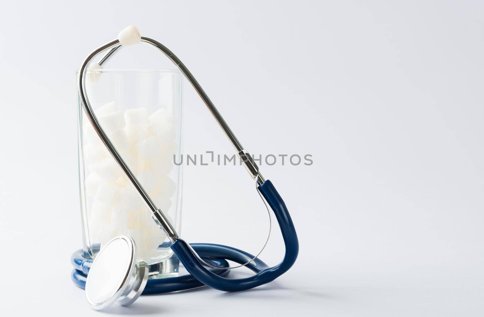 A glass full of white sugar cube sweet food ingredient and doctor stethoscope, studio shot isolated white background, health high blood risk of diabetes and calorie intake concept and unhealthy drink