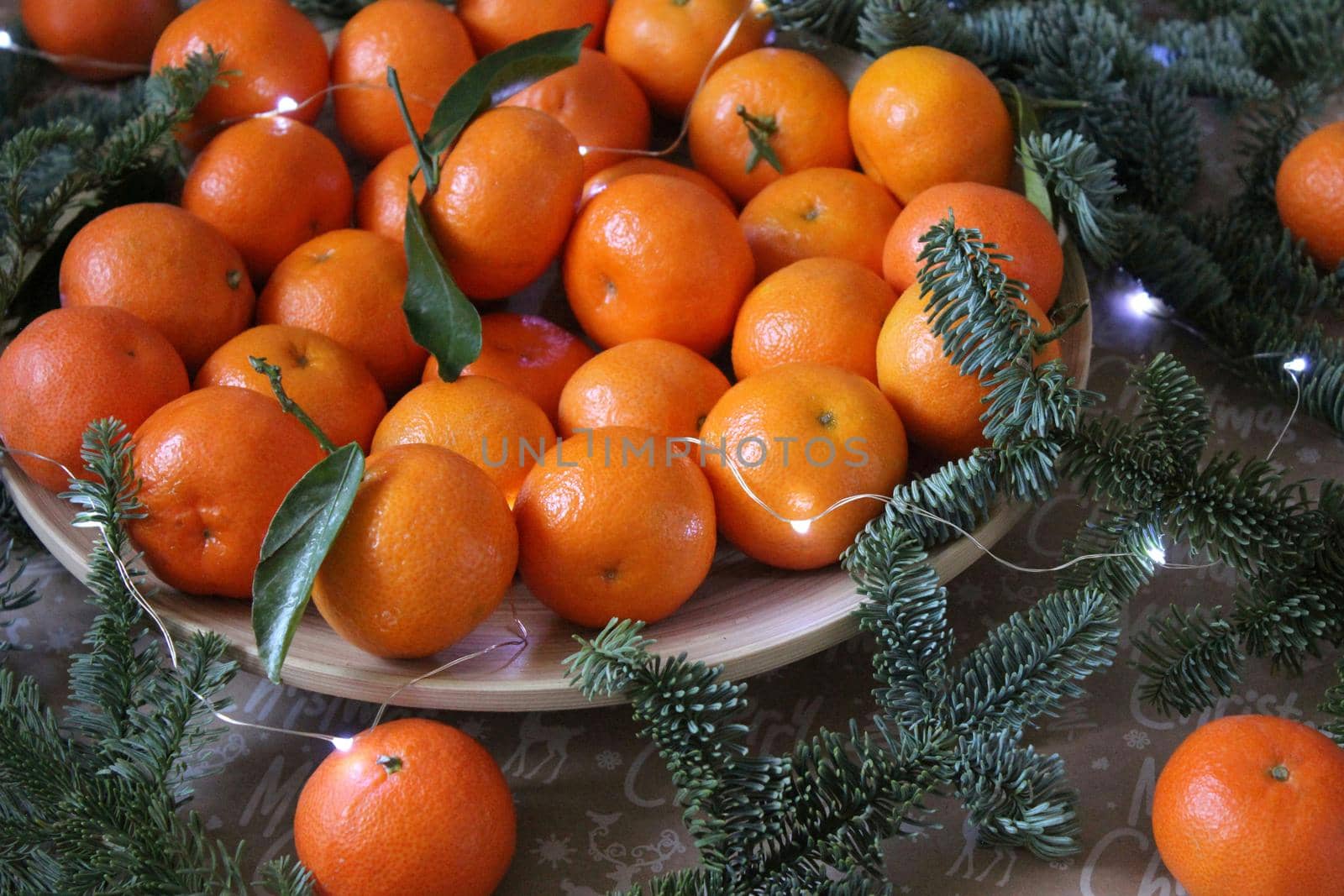 Many small tangerines on the plate are decorated with a garland and coniferous green branches. Texture or background
