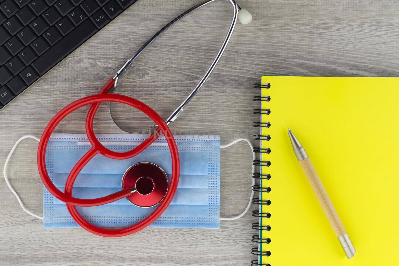 Coronavirus or Covid-19 medical concept with a colorful red stethoscope coiled over a surgical face mask alongside a notebook and pen on a wooden desk with computer keyboard viewed top down