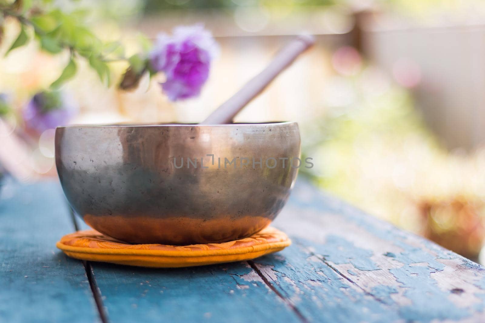 Metal singing bowl on a rustic green, wooden table outdoors. Flowers in the colourful, blurry background