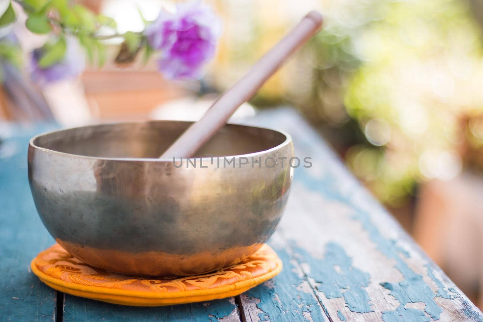 Metal singing bowl on a rustic green, wooden table outdoors. Flowers in the colourful, blurry background