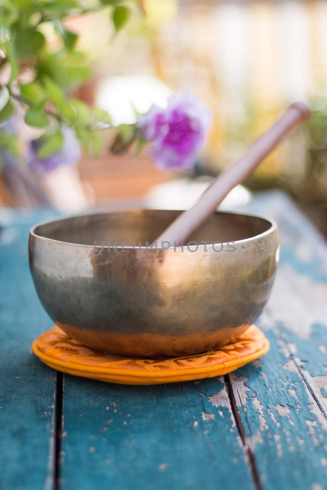 Metal singing bowl on a rustic green, wooden table outdoors. Flowers in the colourful, blurry background