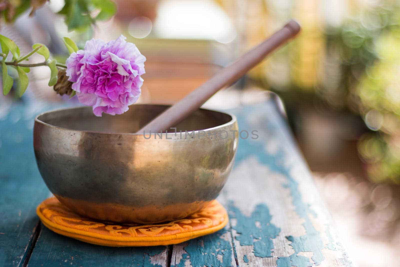 Metal singing bowl on a rustic green, wooden table outdoors. Flowers in the colourful, blurry background