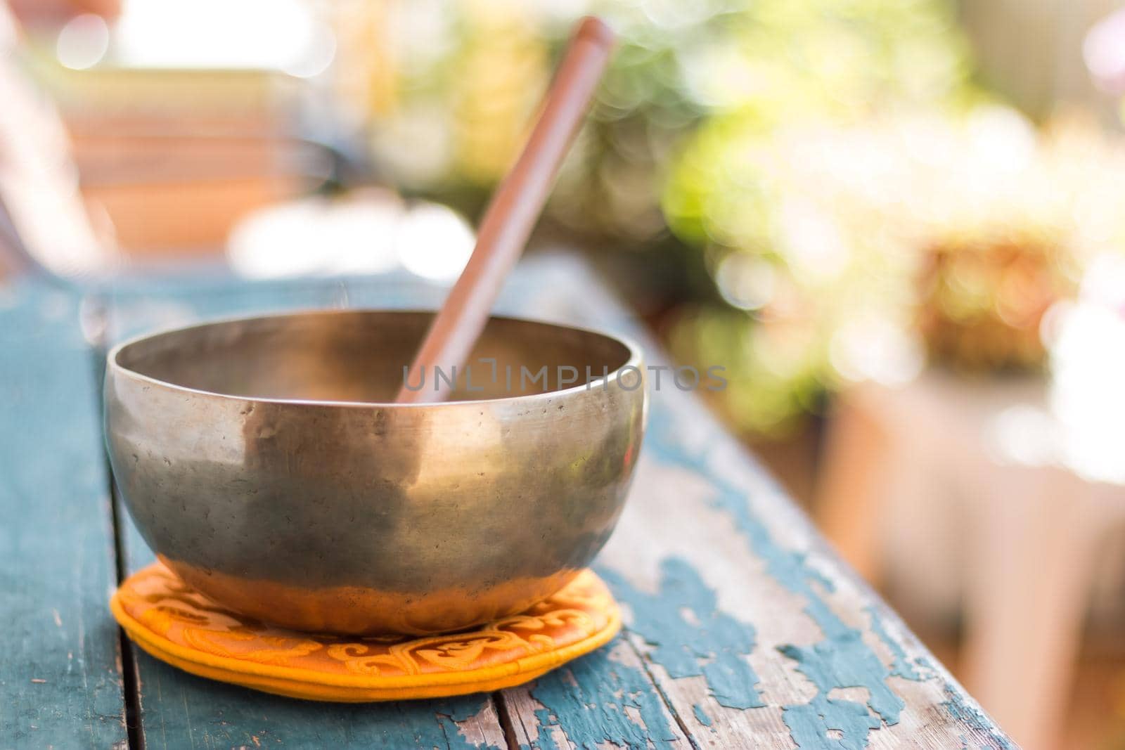 Metal singing bowl on a rustic green, wooden table outdoors. Flowers in the colourful, blurry background