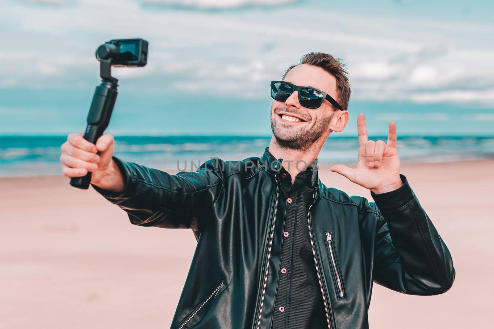 Youthful Blogger in Sunglasses Making Selfie or Streaming Video at the Beach Using Action Camera with Gimbal Camera Stabilizer. Handsome Man in Black Clothes Making Photo Against the Sea