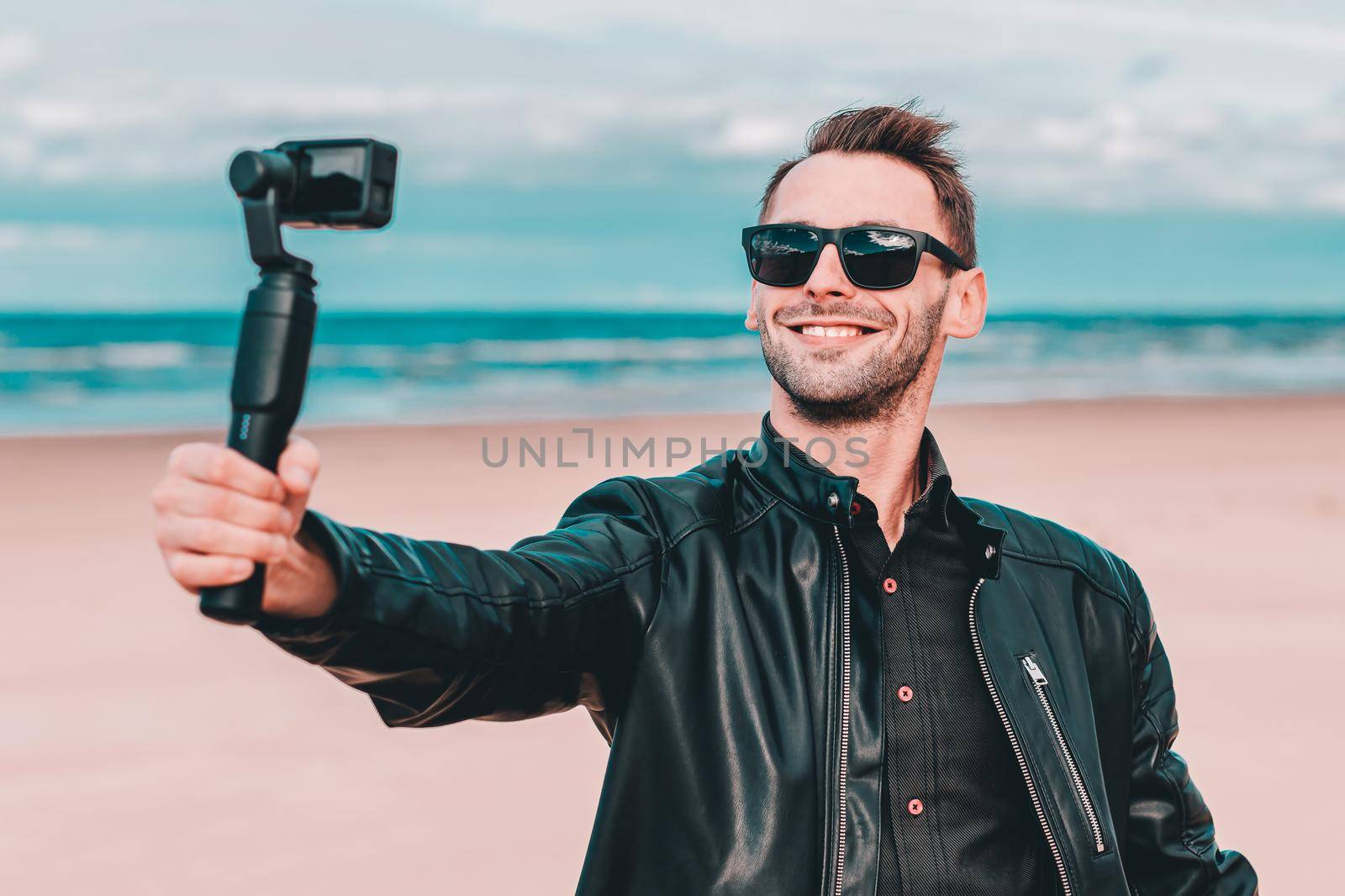Portrait of Smiling Blogger in Sunglasses Making Selfie or Streaming Video at the Beach Using Action Camera with Gimbal Camera Stabilizer. Man in Black Clothes Making Photos