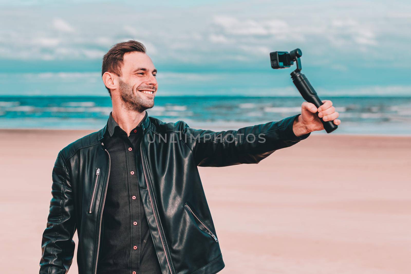 Smiling Young Blogger Making Selfie or Streaming Video at the Beach Using Action Camera with Gimbal Camera Stabilizer. Man in Black Clothes Making Photo Against the Sea