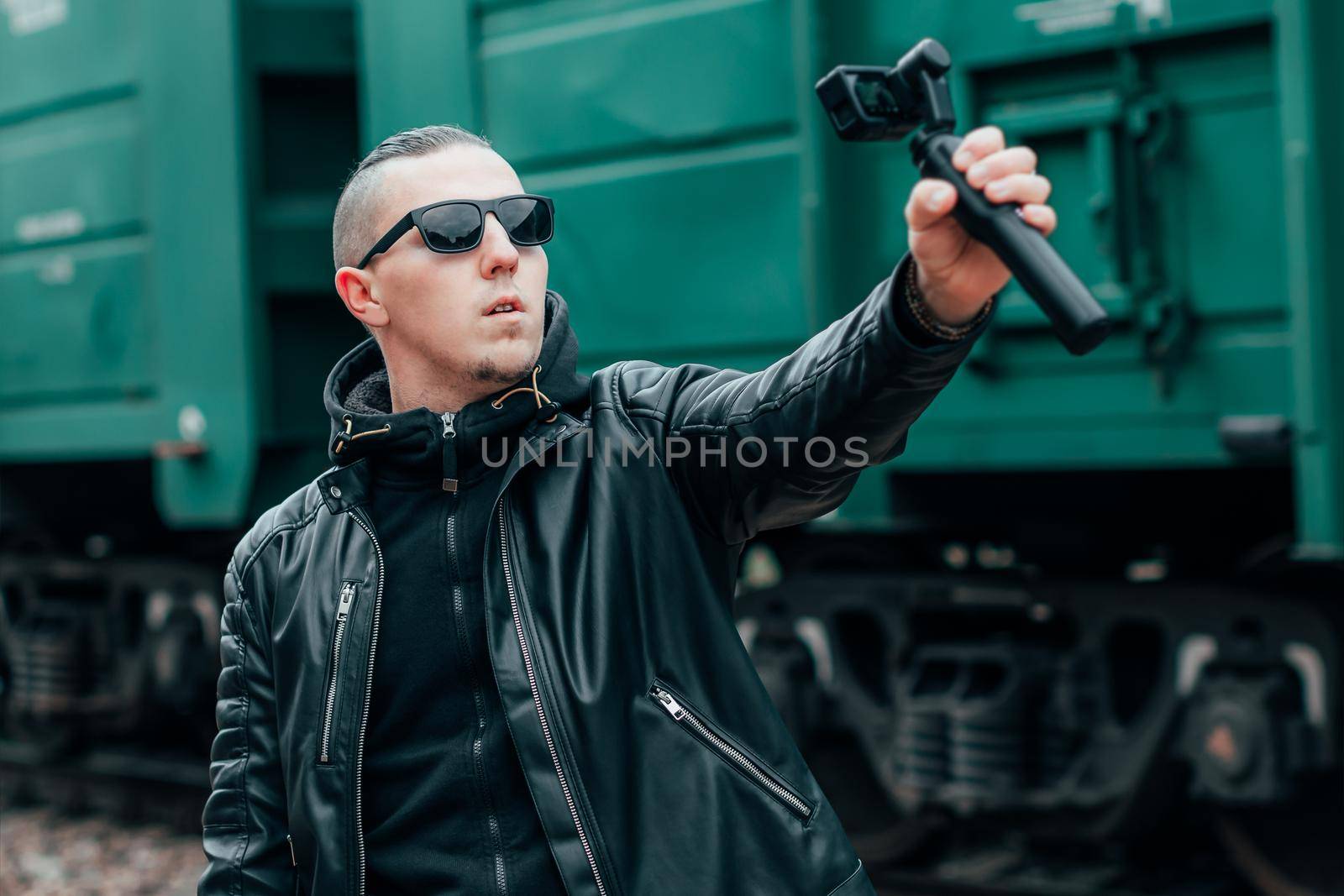 Handsome Guy in Black Clothes and Sunglasses Making Selfie or Streaming Video Using Action Camera with Gimbal Camera Stabilizer at Railway