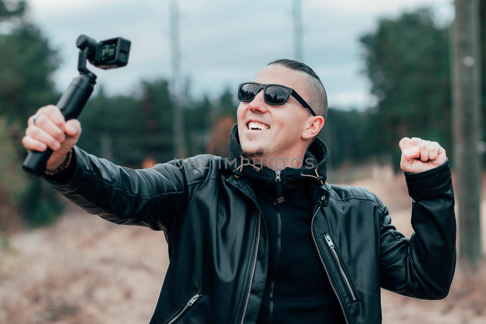 Blogger in Sunglasses Making Selfie or Streaming Video at the Pine Forest Using Action Camera with Gimbal Camera Stabilizer. Handsome Man in Black Clothes Making Photo