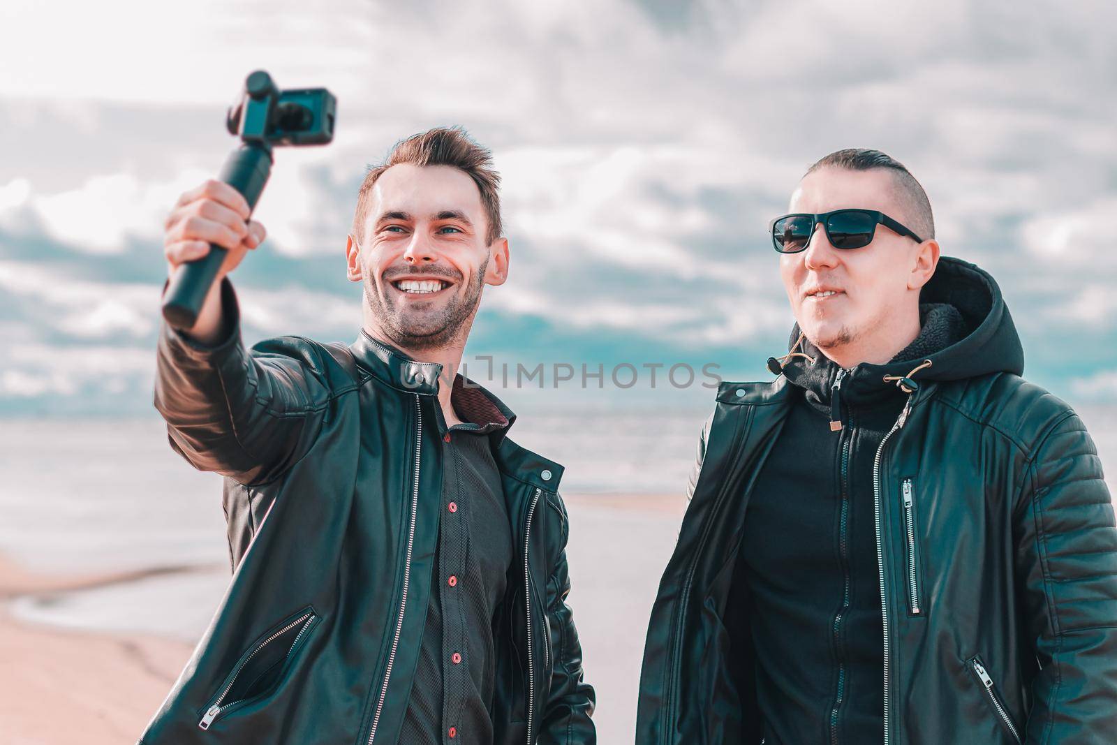 Two Handsome Smiling Friends Making Selfie Using Action Camera with Gimbal Stabilizer at the Beach. Youthful Men in Black Clothes Having Fun by Making Photos