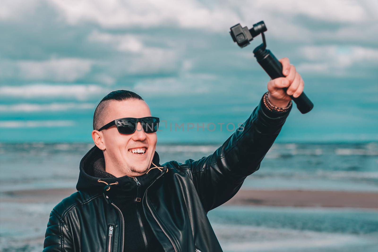 Smiling Young Male Blogger in Sunglasses Making Selfie or Streaming Video at the Beach Using Action Camera with Gimbal Camera Stabilizer. Hipster in Black Clothes Making Photo Against the Sea