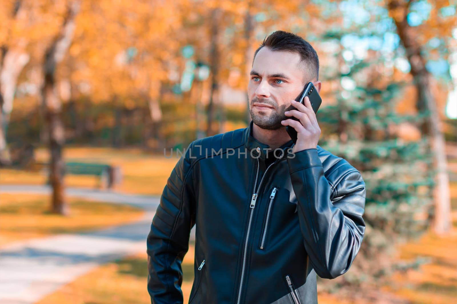 Handsome Young Man Talking on the Phone at the Beautiful Autumn Park. Youthful Guy Using Smartphone for the Call Outdoors at Sunny Day