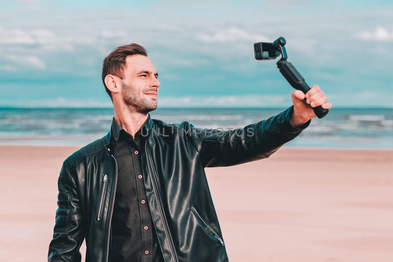 Youthful Blogger Making Selfie or Streaming Video at the Beach Using Action Camera with Gimbal Camera Stabilizer. Handsome Guy in Black Clothes Making Photo Against the Sea