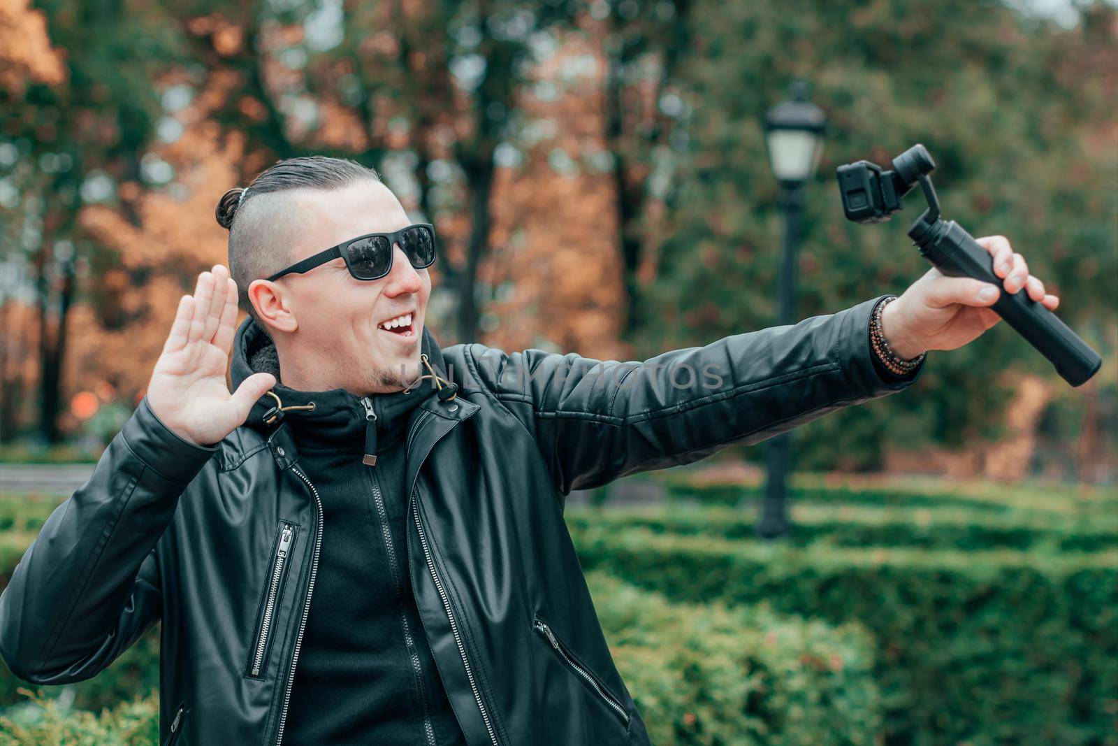 Blogger in Sunglasses Making Selfie or Streaming Video at the Autumn Park Using Action Camera with Gimbal Camera Stabilizer. Handsome Man in Black Clothes Making Photo