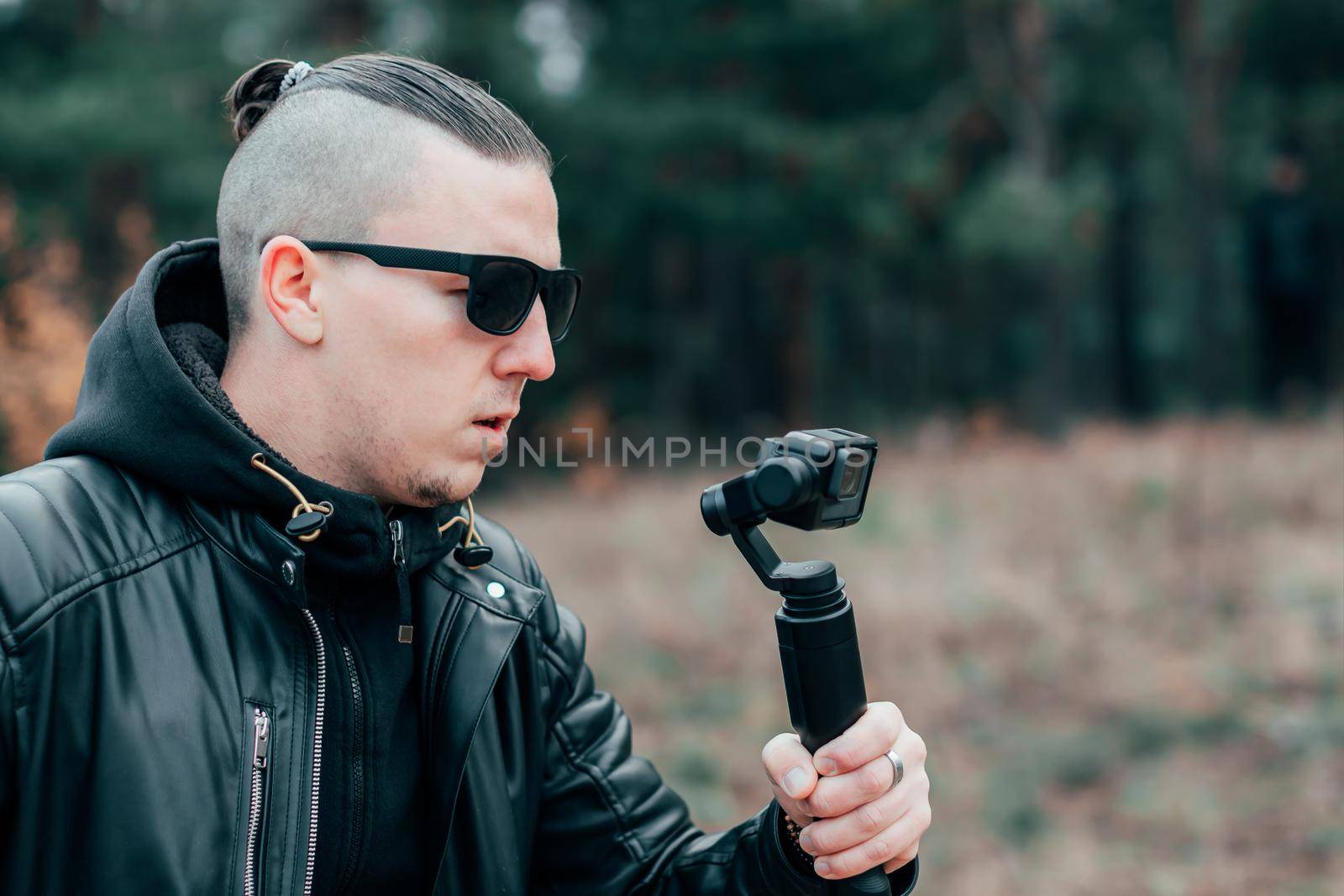 Youthful Blogger in Black Clothes and Sunglasses Making Video Using Action Camera with Gimbal Camera Stabilizer in Pine Forest