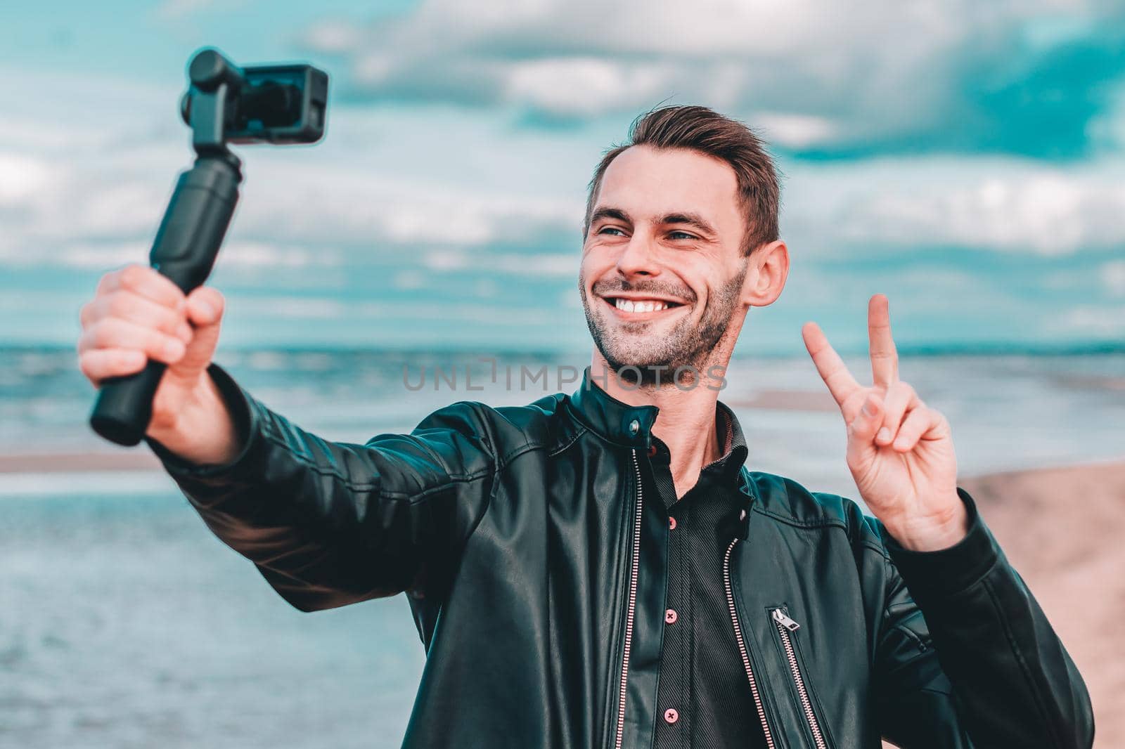 Smiling Young Blogger Making Selfie or Streaming Video at the Beach Using Action Camera with Gimbal Camera Stabilizer. Man in Black Clothes Making Photo Showing Peace