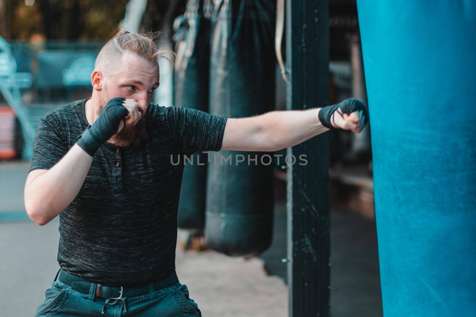 Street Fighter in Black Clothes and Bandages on the Wrist Boxing in Punching Bag Outdoors. Young Man Doing Box Training and Practicing His Punches at the Outside Gym