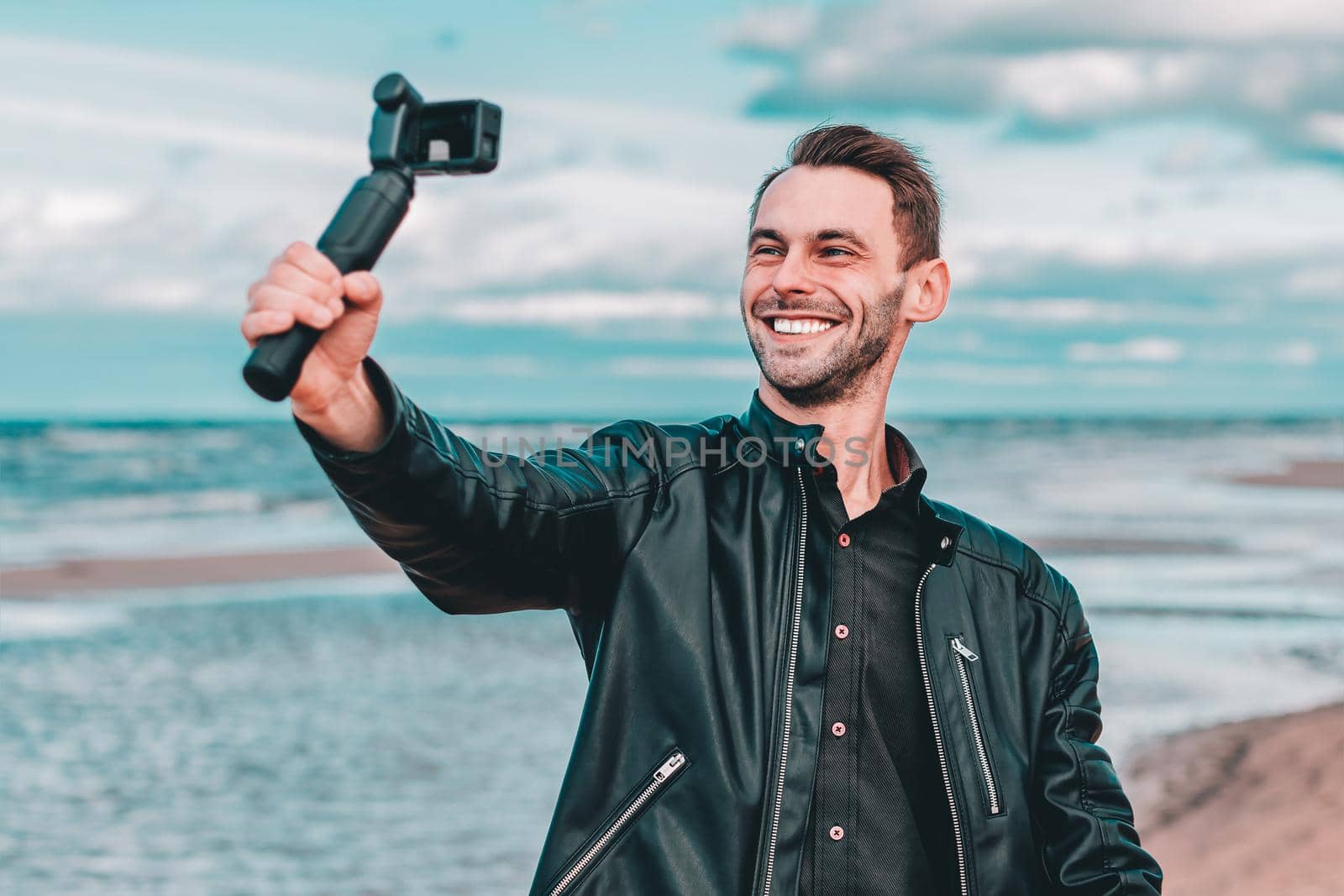 Smiling Young Male Blogger Making Selfie or Streaming Video at the Beach Using Action Camera with Gimbal Camera Stabilizer. Man in Black Clothes Making Photo Against the Sea