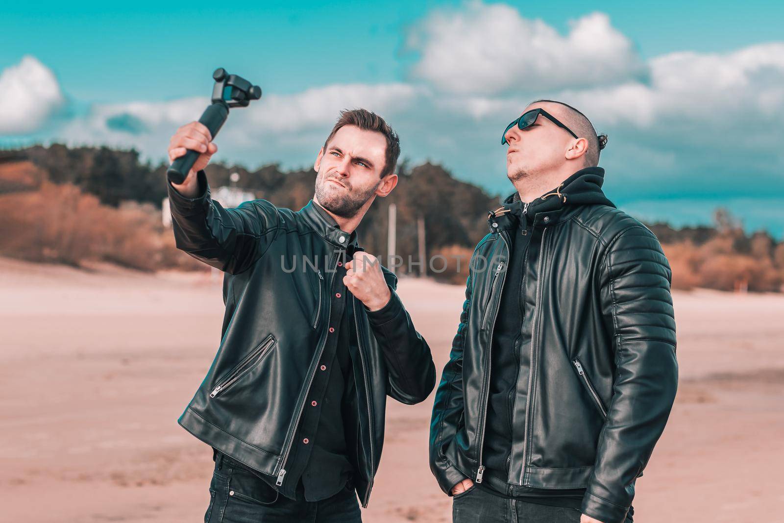 Two Handsome Male Friends Making Selfie Using Action Camera with Gimbal Stabilizer at the Beach. Youthful Men in Black Clothes Having Fun by Making Photos