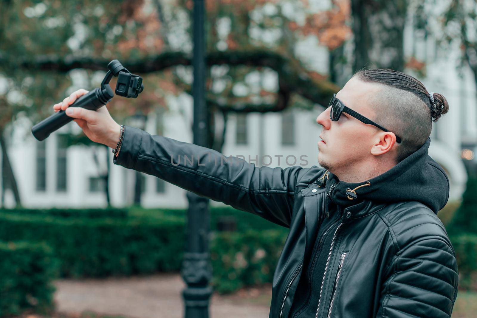 Blogger in Sunglasses Making Selfie or Streaming Video at the Autumn Park Using Action Camera with Gimbal Camera Stabilizer. Handsome Man in Black Clothes Making Photo