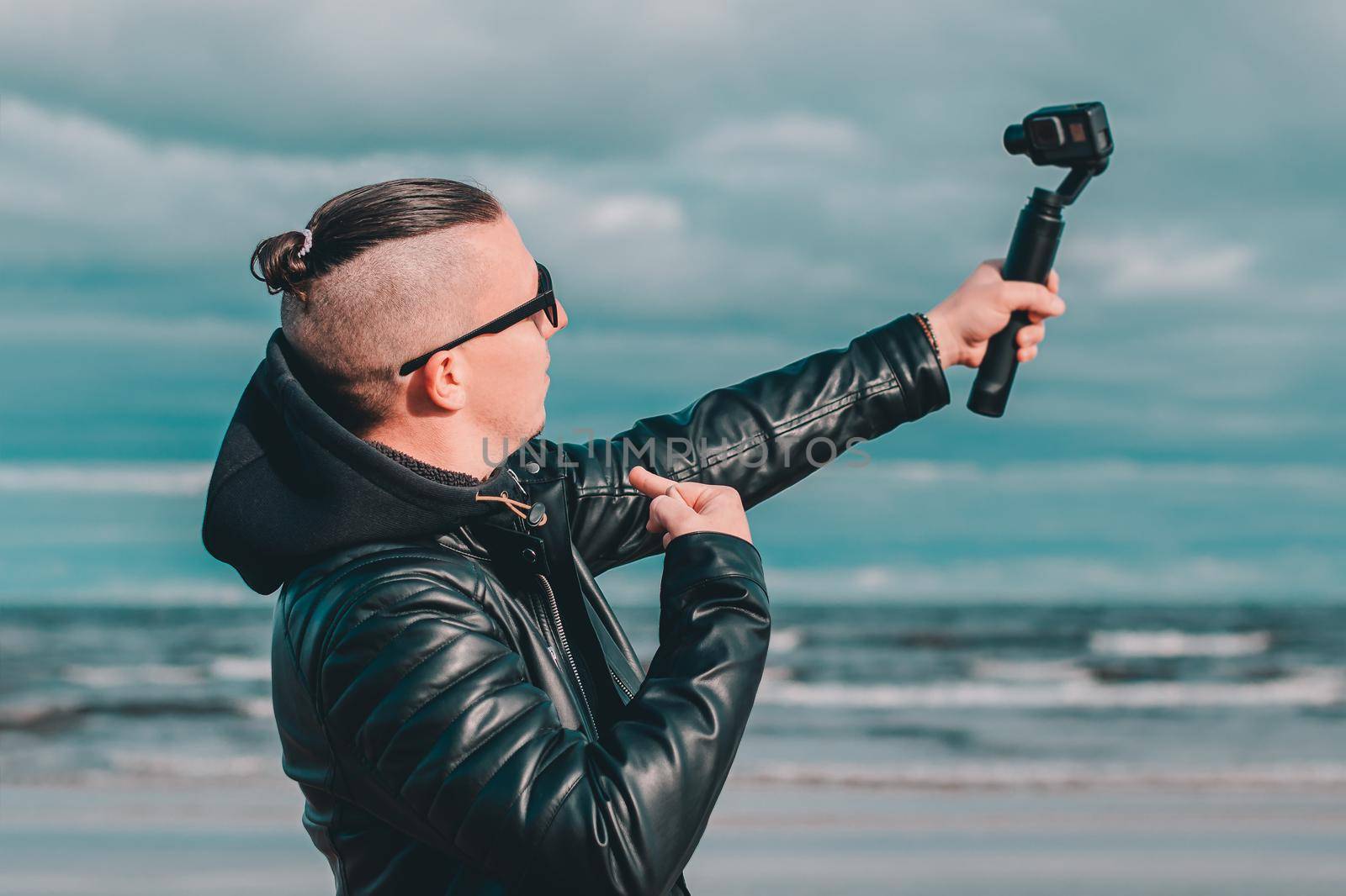 Young Blogger in Sunglasses Making Selfie or Streaming Video at the Beach Using Action Camera with Gimbal Camera Stabilizer. Hipster in Black Clothes Making Photo Against the Sea