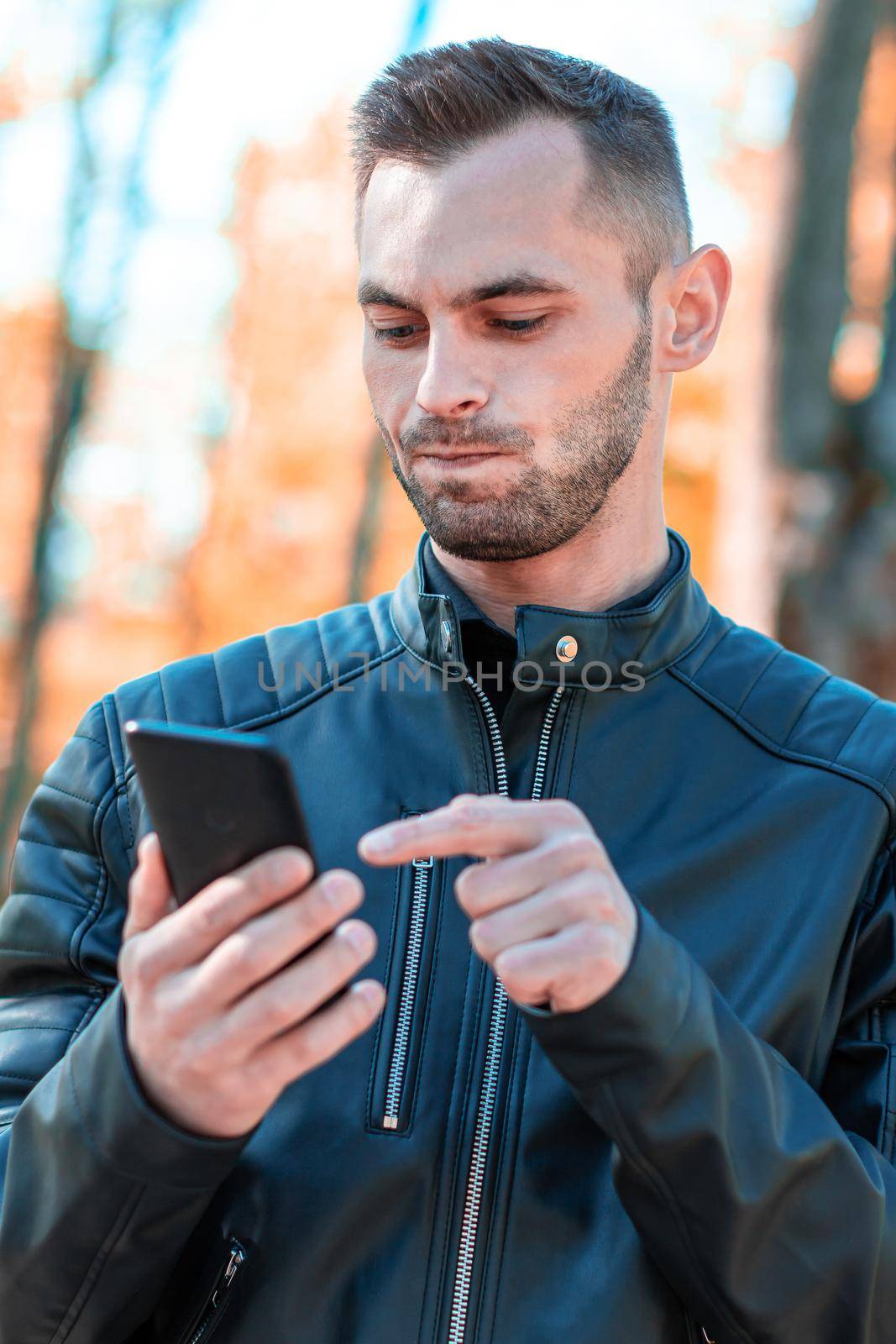 Youthful Guy Using Black Smartphone at the Beautiful Autumn Park. Handsome Young Man with Mobile Phone at Sunny Day - Medium CloseUp Shot