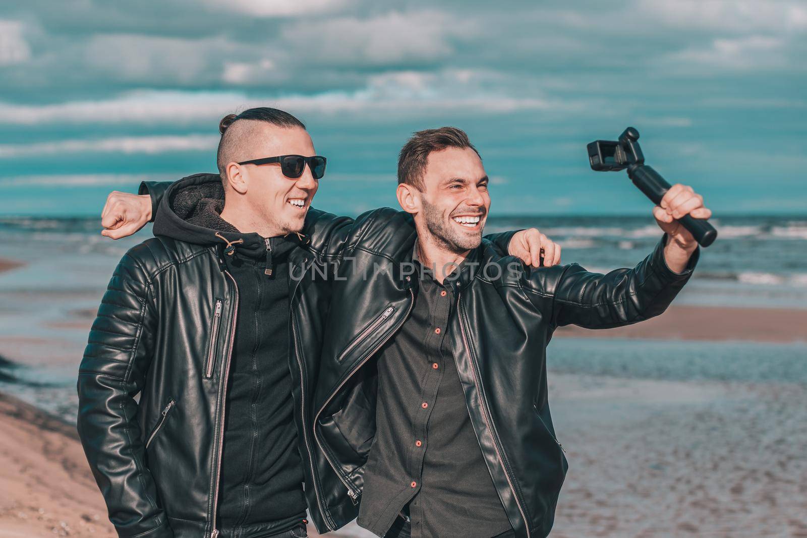 Two Handsome Smiling Friends Making Selfie Using Action Camera with Gimbal Stabilizer at the Beach. Youthful Men in Black Clothes Having Fun by Making Photos