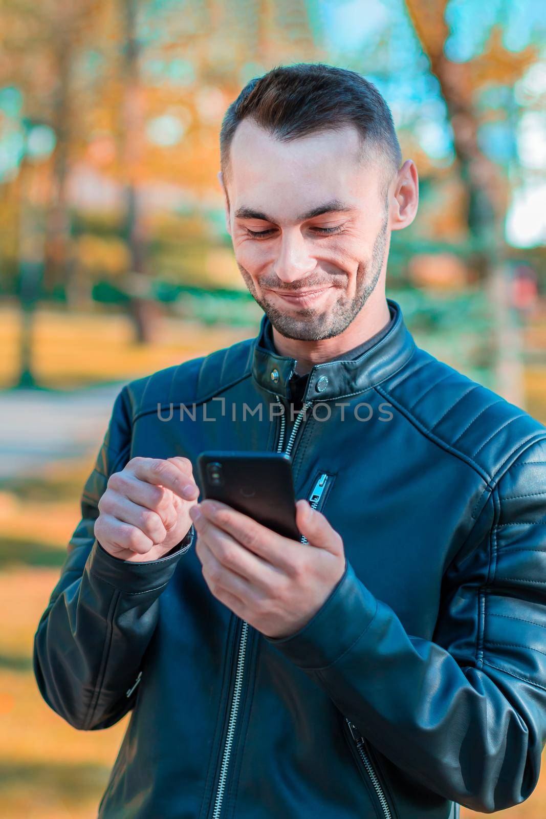 Youthful Satisfied Guy Using Black Smartphone at the Beautiful Autumn Park. Handsome Smiling Young Man with Mobile Phone at Sunny Day - Medium CloseUp Portrait