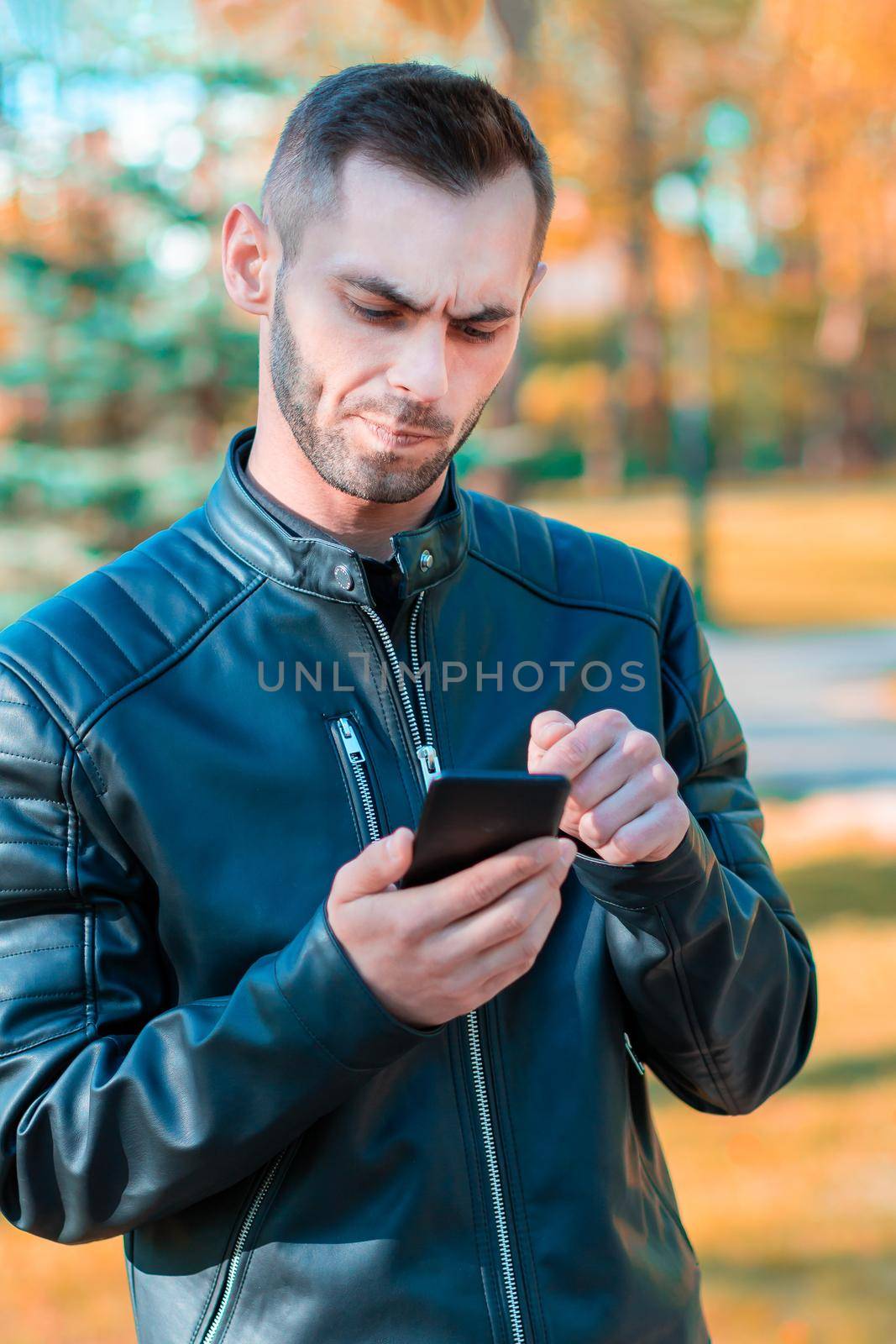 Youthful Frowning Guy Using Black Smartphone at the Beautiful Autumn Park. Handsome Young Man with Mobile Phone at Sunny Day - Medium Shot Portrait