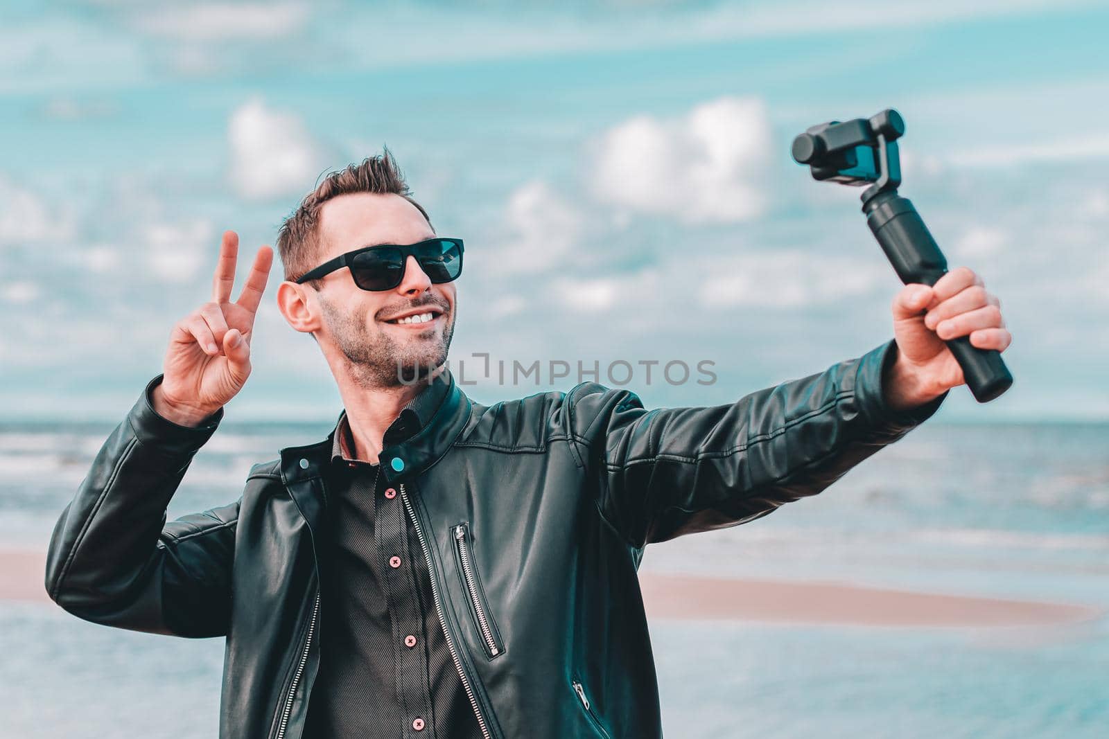 Youthful Blogger in Sunglasses Making Selfie or Streaming Video at the Beach Using Action Camera with Gimbal Camera Stabilizer. Handsome Man in Black Clothes Making Photo Against the Sea