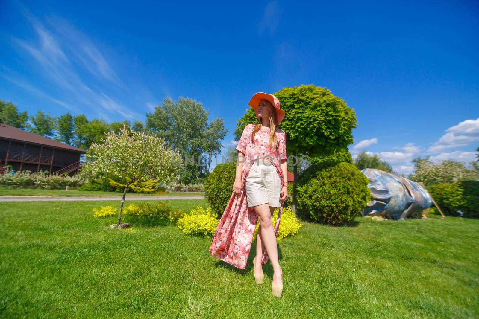 A charming girl in a light summer sundress and a pareo hat is walking in a green park. Enjoys warm sunny summer days.